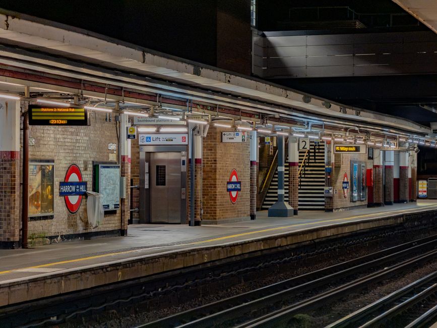 Nighttime view of the Harrow & Wealdstone London Underground station platform, featuring brick walls with signs and posters, illuminated by overhead lighting. Central to the scene are two stainless st