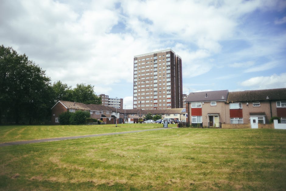 A residential area featuring a small grassy field in the foreground with a narrow footpath running across it. In the middle ground, there are several low-rise houses with brown brick walls, sloped til