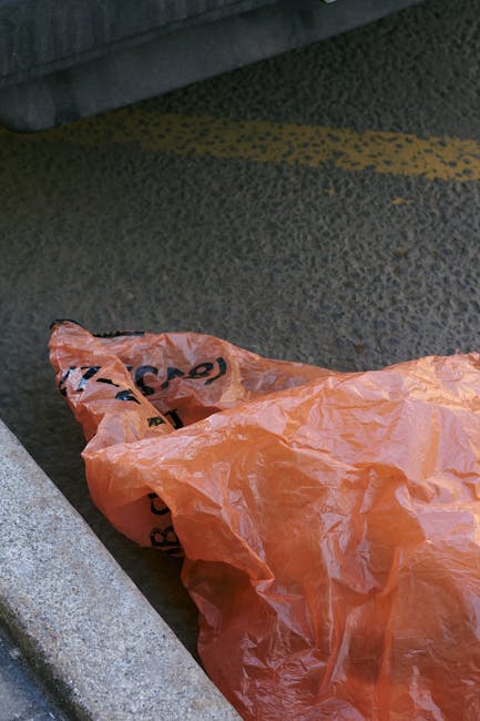 A crumpled orange plastic rubbish bag lying on a textured asphalt surface next to a concrete curb in an outdoor urban environment. The bag appears to contain waste and is partially open, with some bla