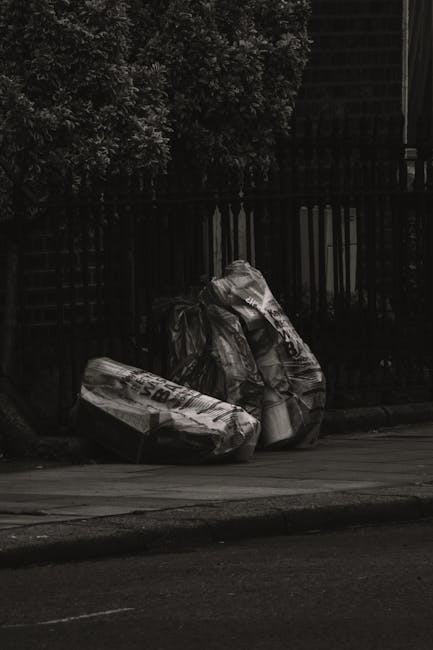 A crumpled orange plastic rubbish bag lying on a textured asphalt surface next to a concrete curb in an outdoor urban environment. The bag appears to contain waste and is partially open, with some bla